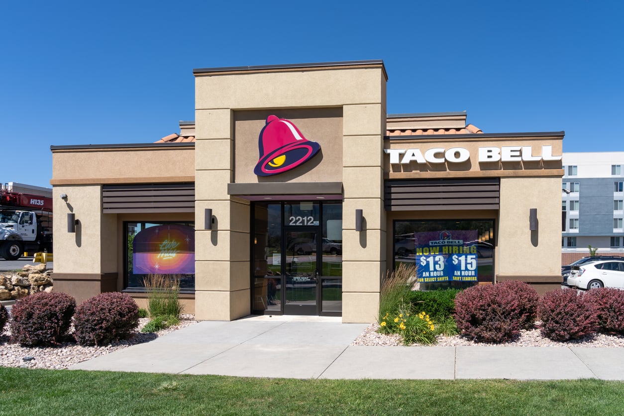 A Taco Bell restaurant with a large logo above the entrance and hiring signs on the windows, surrounded by landscaping and a clear blue sky.