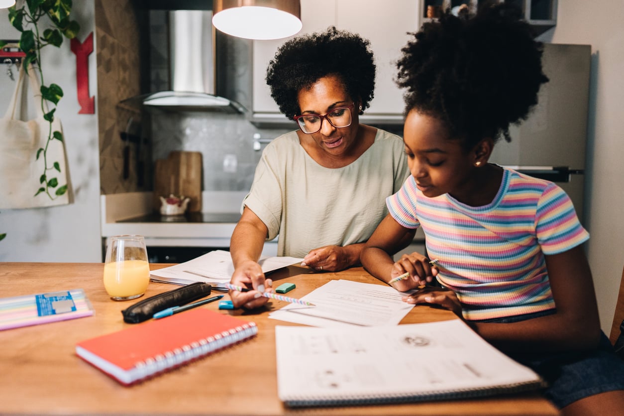 A woman and a girl sit at a kitchen table, working on homework together. Surrounded by notebooks, papers, and orange juice, they appear focused—reflecting how families in America are having fewer children but cherishing quality time together.