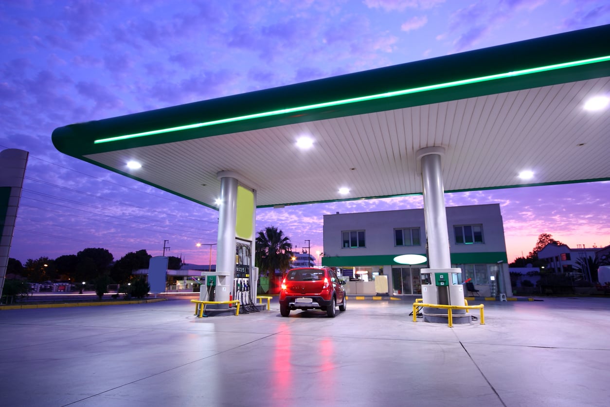 A red SUV refuels at a modern gas station under a large canopy at dusk, perhaps making a quick stop to tick off items on the road trip checklist. The sky glows purple, and the station is mostly empty, with buildings and palm trees silhouetted in the background.