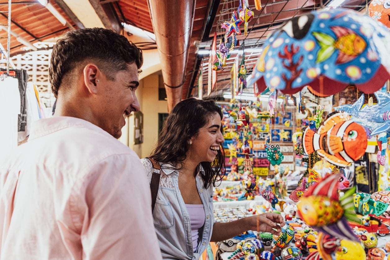 A man and woman smile and look at colorful fish-shaped crafts and souvenirs at an outdoor market stall. Bright decorations hang around them in a lively, cheerful setting.