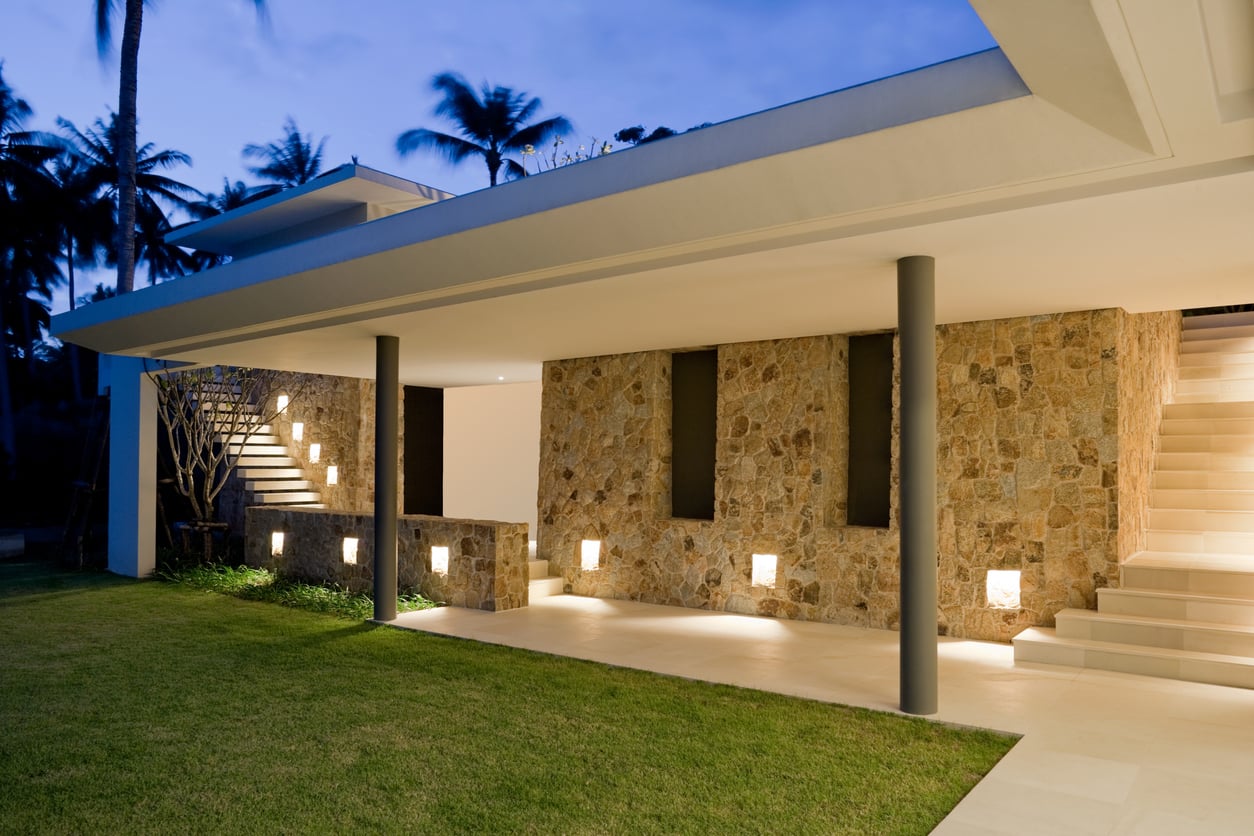 Modern house exterior at dusk with stone walls, tall vertical windows, and a flat roof. Outdoor lighting illuminates the steps and walkway, with a green lawn in the foreground and palm trees in the background.