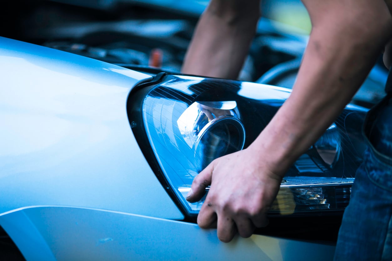A person’s hands work on the front headlight of a silver car, with the hood open. The focus is on the headlight and arms, highlighting practical car maintenance hacks during routine repair.