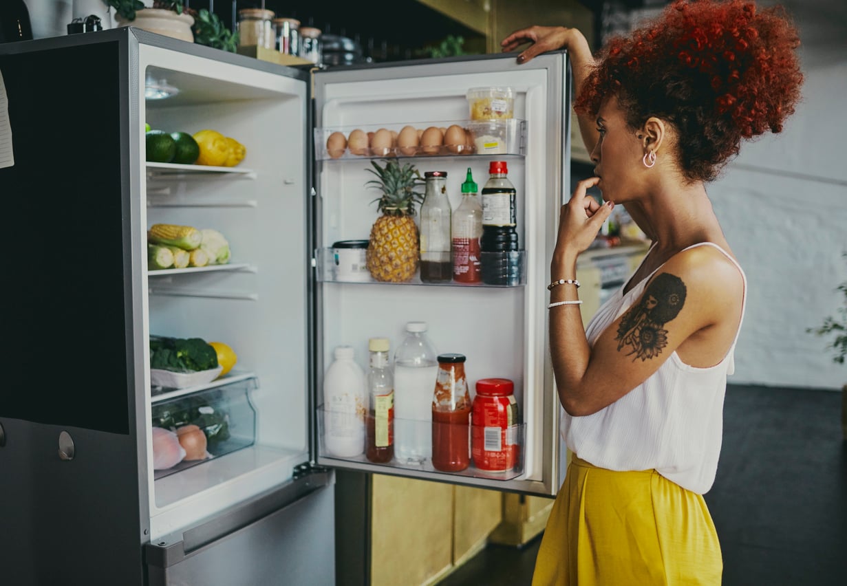 A woman with curly red hair and a sun and moon tattoo stands thoughtfully in front of an open refrigerator filled with eggs, fruit, vegetables, and condiments, reflecting how women are changing their spending behaviors by making mindful choices about what they purchase.