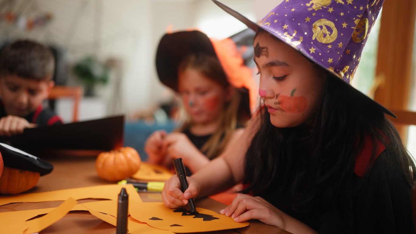 Children in Halloween costumes enjoy festive Halloween activities as they draw jack-o’-lantern faces on orange paper at a table, surrounded by small pumpkins. One child wears a purple witch hat and has face paint.