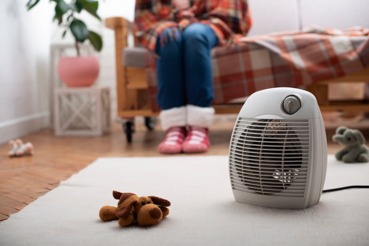 A portable electric space heater sits on a carpeted floor with a toy dog nearby. In the background, a person with pink slippers and a blanket over their lap sits on a sofa.