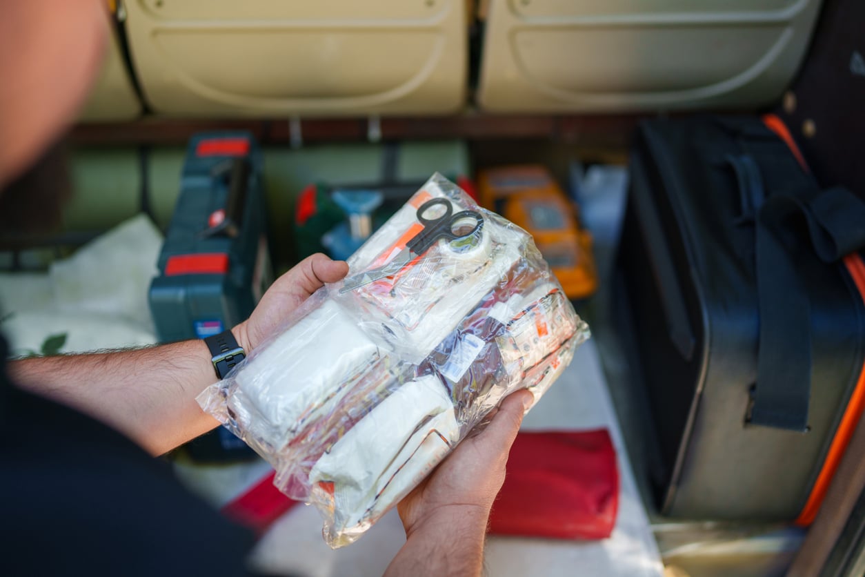 A person holds a clear plastic bag containing medical supplies, such as bandages and scissors—essential items on any road trip checklist—with various bags and equipment visible in the background.