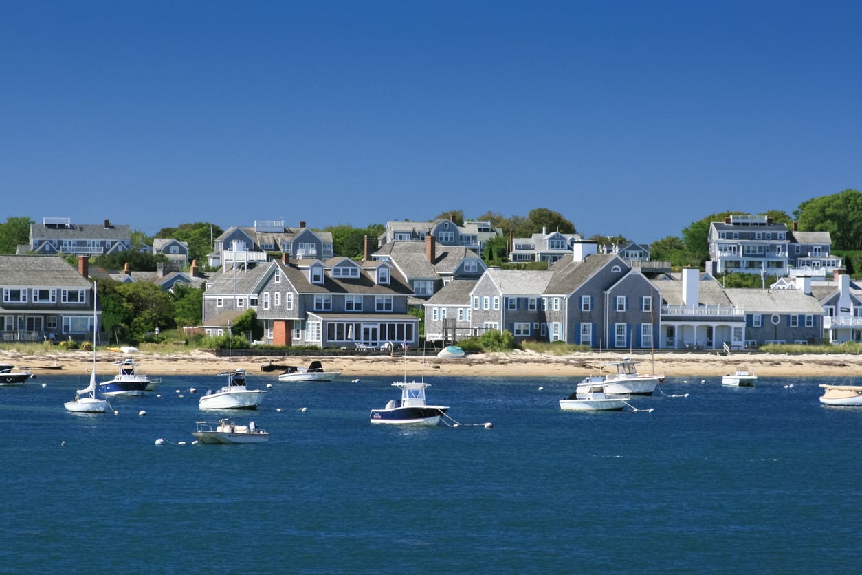 Boats float on blue water in front of a shoreline lined with gray-shingled houses and green trees under a clear blue sky, creating the perfect scene for one of the region’s charming travel destinations.
