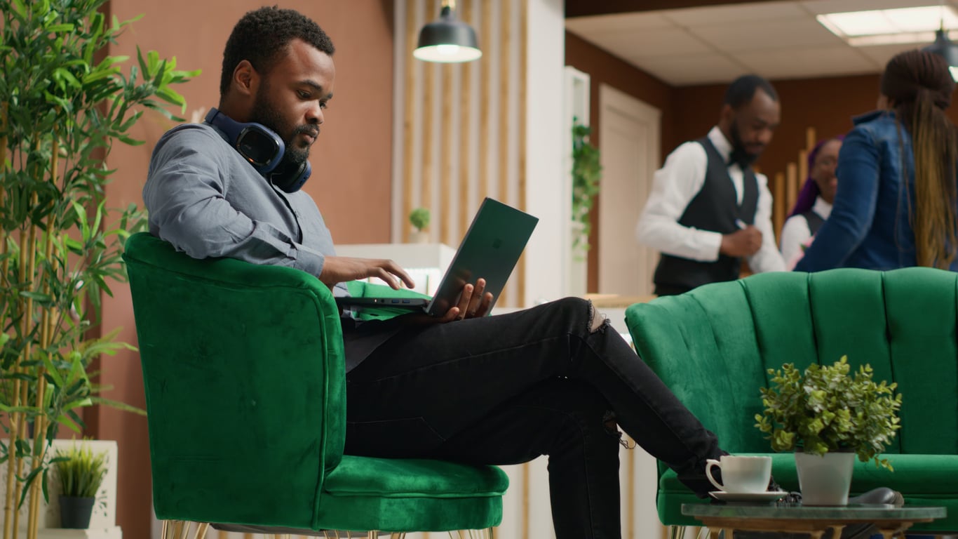 A man sits on a green chair with headphones around his neck, working on a laptop in a modern, stylish office or cafe. As hotel changes are discussed nearby, a coffee cup and small plant rest on the table beside him while people converse in the background.