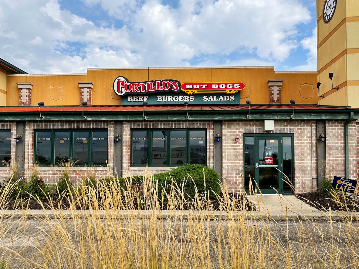 Portillo’s restaurant exterior, one of the iconic regional fast-food chains, features a sign with “Hot Dogs, Beef, Burgers, Salads.” Decorative grasses are in the foreground and a partly cloudy sky is visible above the building.