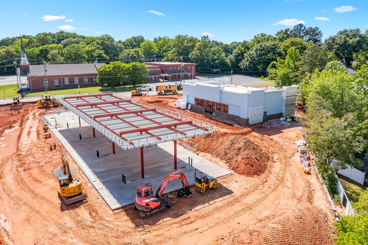 A construction site with steel framework for a building, surrounded by dirt, construction vehicles, trees, and a brick church in the background under a blue sky—nearby, the presence of regional fast-food chains adds a touch of local flavor to the bustling scene.