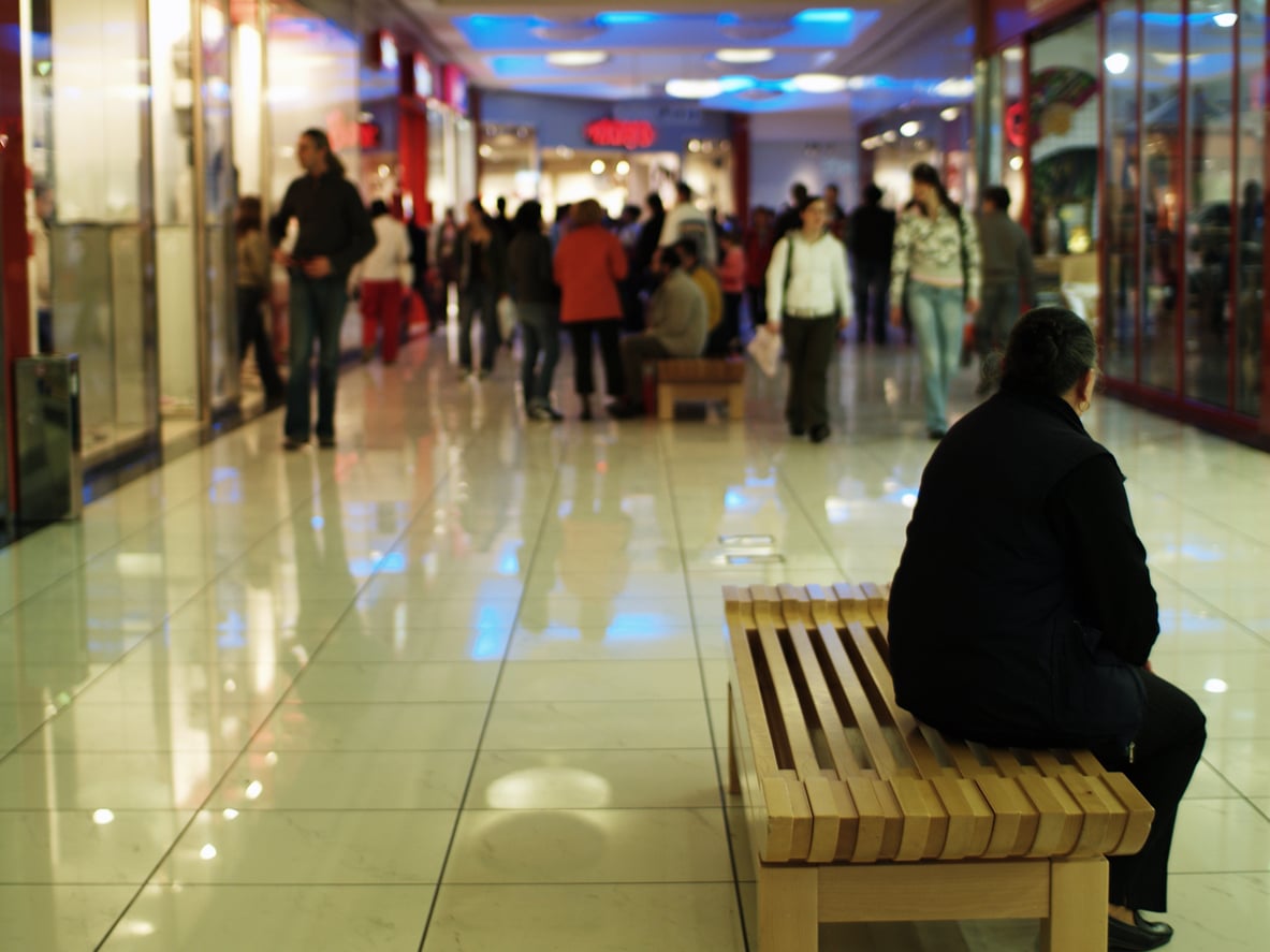 A person sits alone on a wooden bench in a busy shopping mall, facing away. Shiny floors and storefronts reflect lights as people walk in the background.