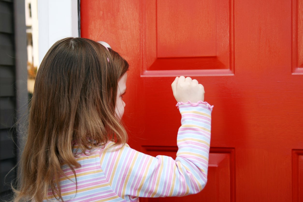 A young girl with long brown hair in a striped shirt is seen from behind as she knocks on a bright red door.