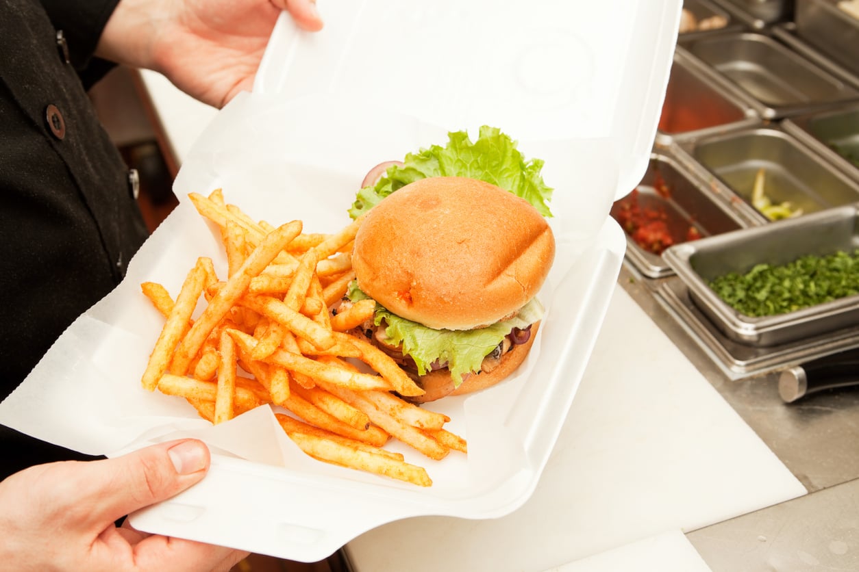 A person holds a takeout container with a hamburger topped with lettuce and a serving of French fries beside it. Containers of toppings are visible in the background.