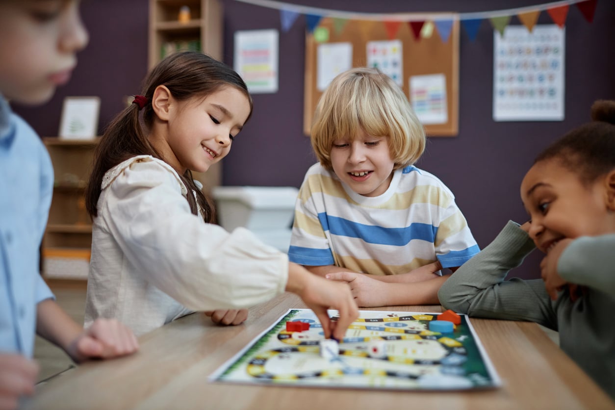 Four children sit around a table, smiling and playing a colorful board game together in a classroom decorated with posters and bunting.