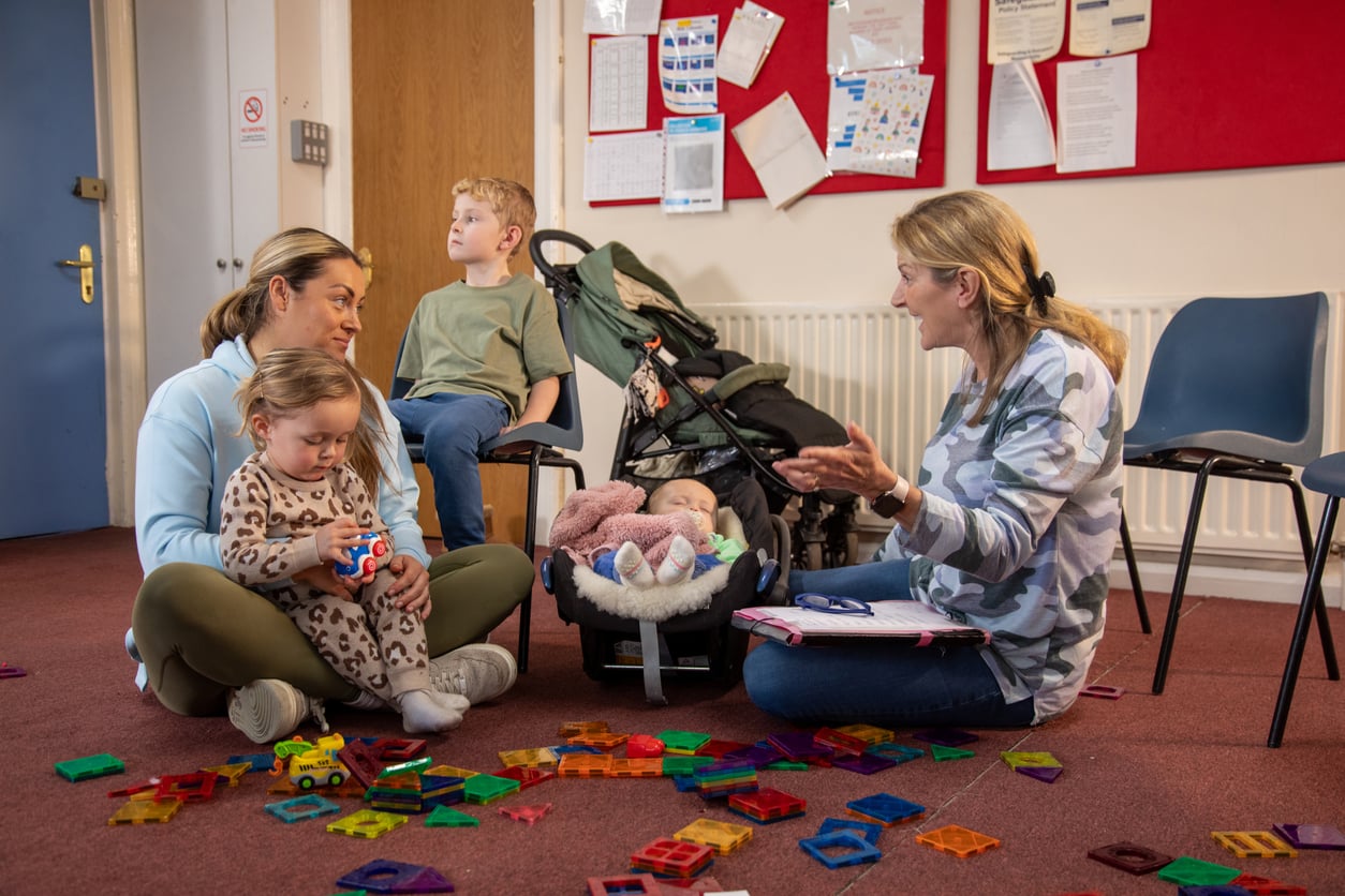 Two women sit on the floor with three young children in a room, showcasing how Families in America Are Having Fewer Children. Colorful toys are scattered around as the women talk and the children play. Noticeboards and chairs are in the background.
