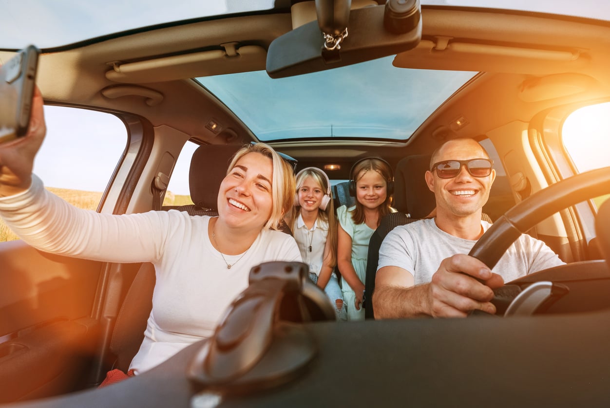 A smiling family of four sits in a car, with a woman taking a selfie, two children in the back seat, and a man driving—all enjoying a sunny day as they practice money saving habits by embarking on an affordable family outing.