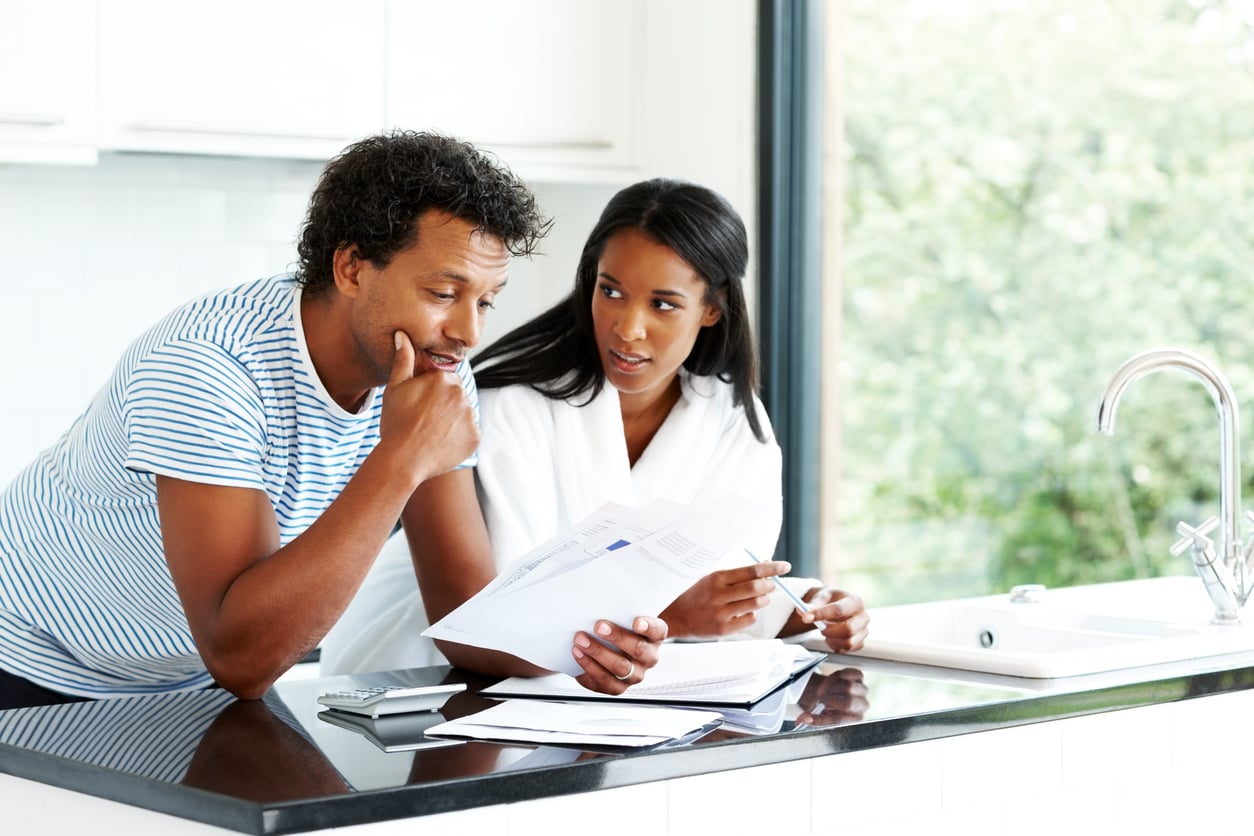 A man and woman stand in a bright kitchen, looking at paperwork together. The man looks thoughtful while holding papers, and the woman sits beside him, pen in hand, appearing concerned. A calculator and notebook are on the counter.