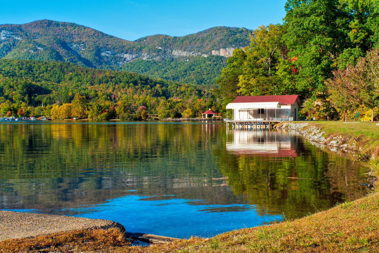 A lake with clear water reflects a house with a red roof, surrounded by trees showing autumn colors, with forested mountains in the background under a clear blue sky.