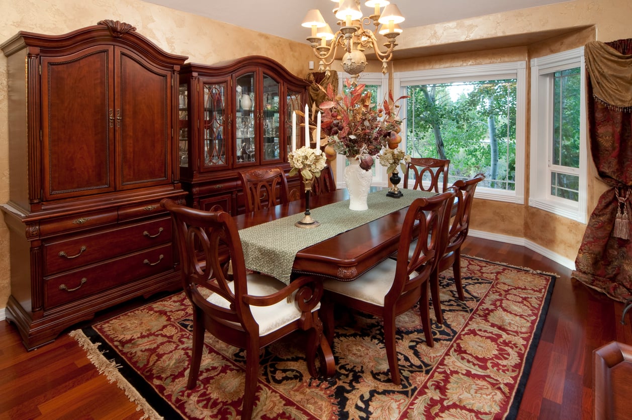 Elegant dining room with wooden table, six chairs, a floral centerpiece, chandelier, china cabinetsāone of the home features homeowners no longer wantābay window, and patterned rug on polished hardwood floors. Large windows overlook greenery outside.