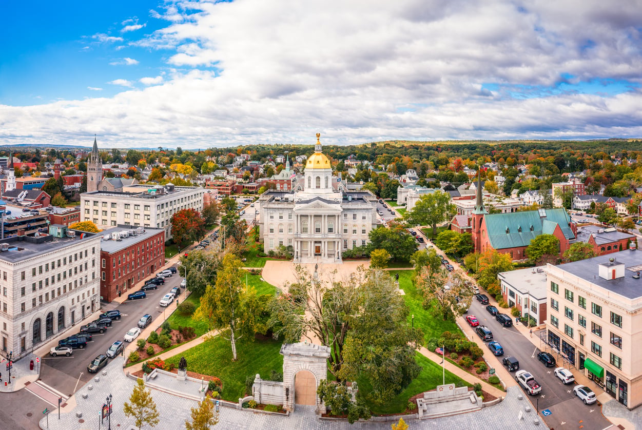 Aerial view of a travel-friendly white capitol building with a golden dome, surrounded by trees with autumn foliage, city streets, cars, and nearby buildings under a partly cloudy sky.