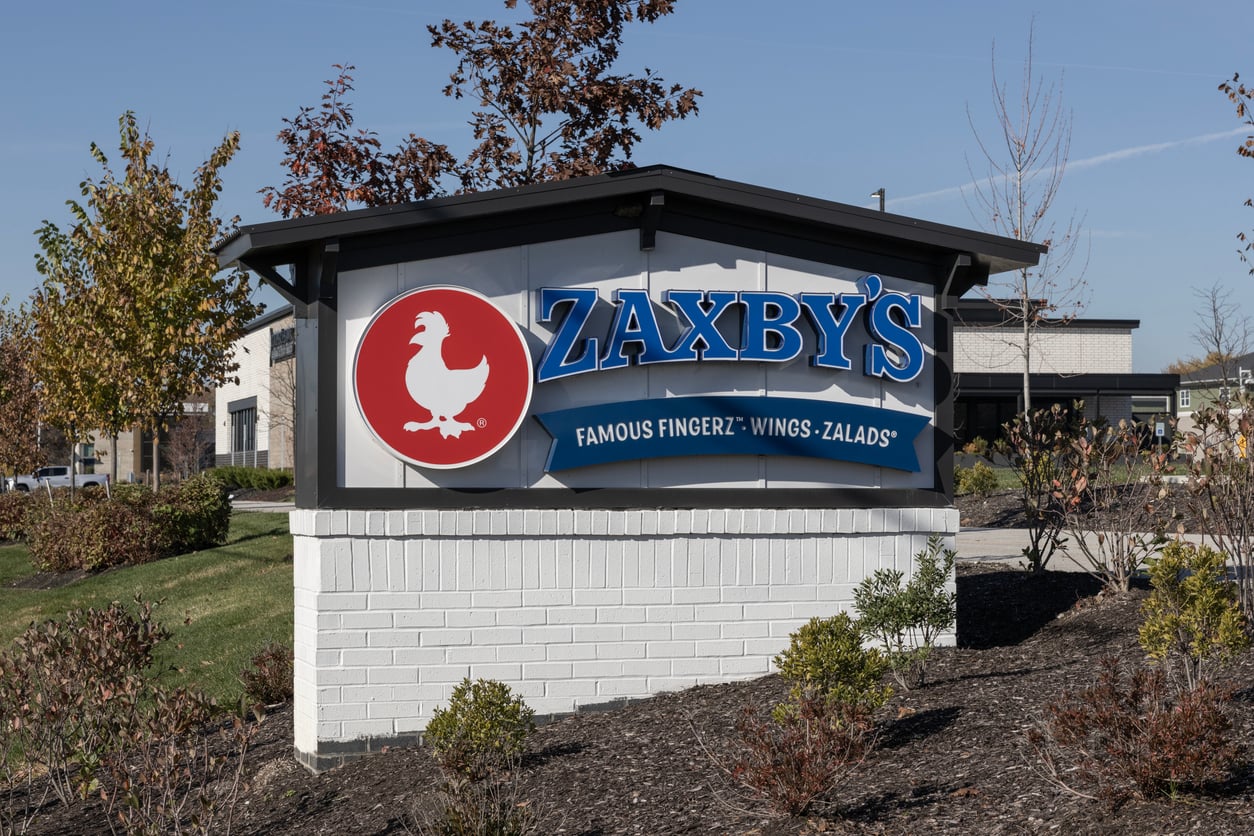 A Zaxby’s restaurant sign with a red chicken logo and the words “ZAXBY’S FAMOUS FINGERZ® · WINGS · ZALADS®” rises from a landscaped area with shrubs and trees, representing one of the leading regional fast-food chains known for its Southern-inspired menu.