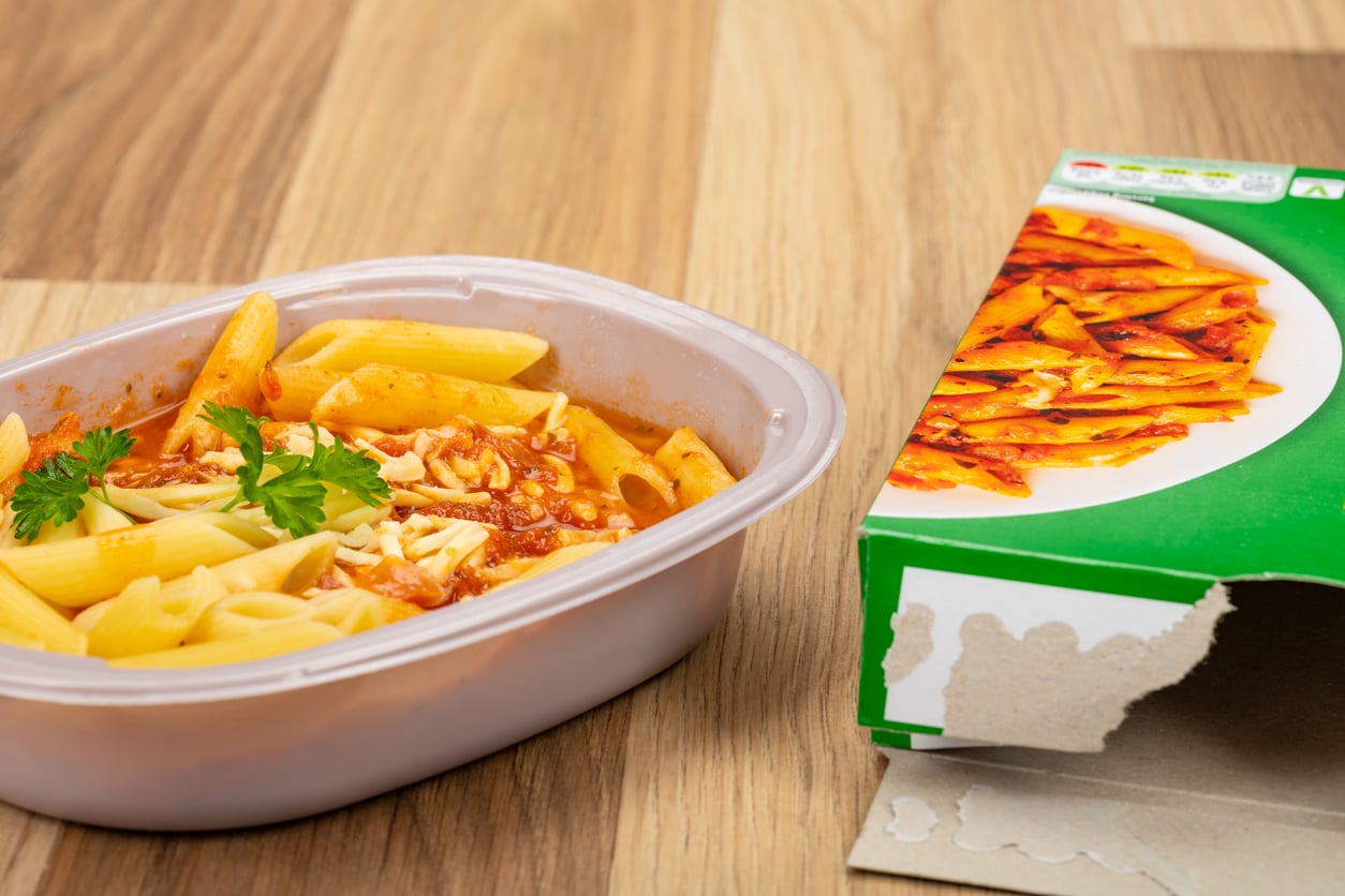 A plastic container of penne pasta with tomato sauce, cheese, and parsley sits next to its green and white cardboard packaging on a wooden table—a delicious example of how money saving habits can make enjoying quality meals both convenient and affordable.