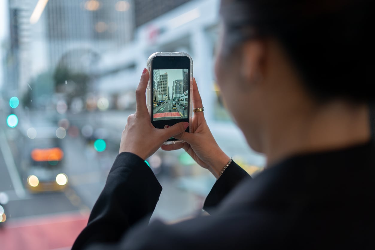 Person taking a photo of a busy city street through a window with a smartphone; buildings, cars, and traffic lights are visible on the phone screen.