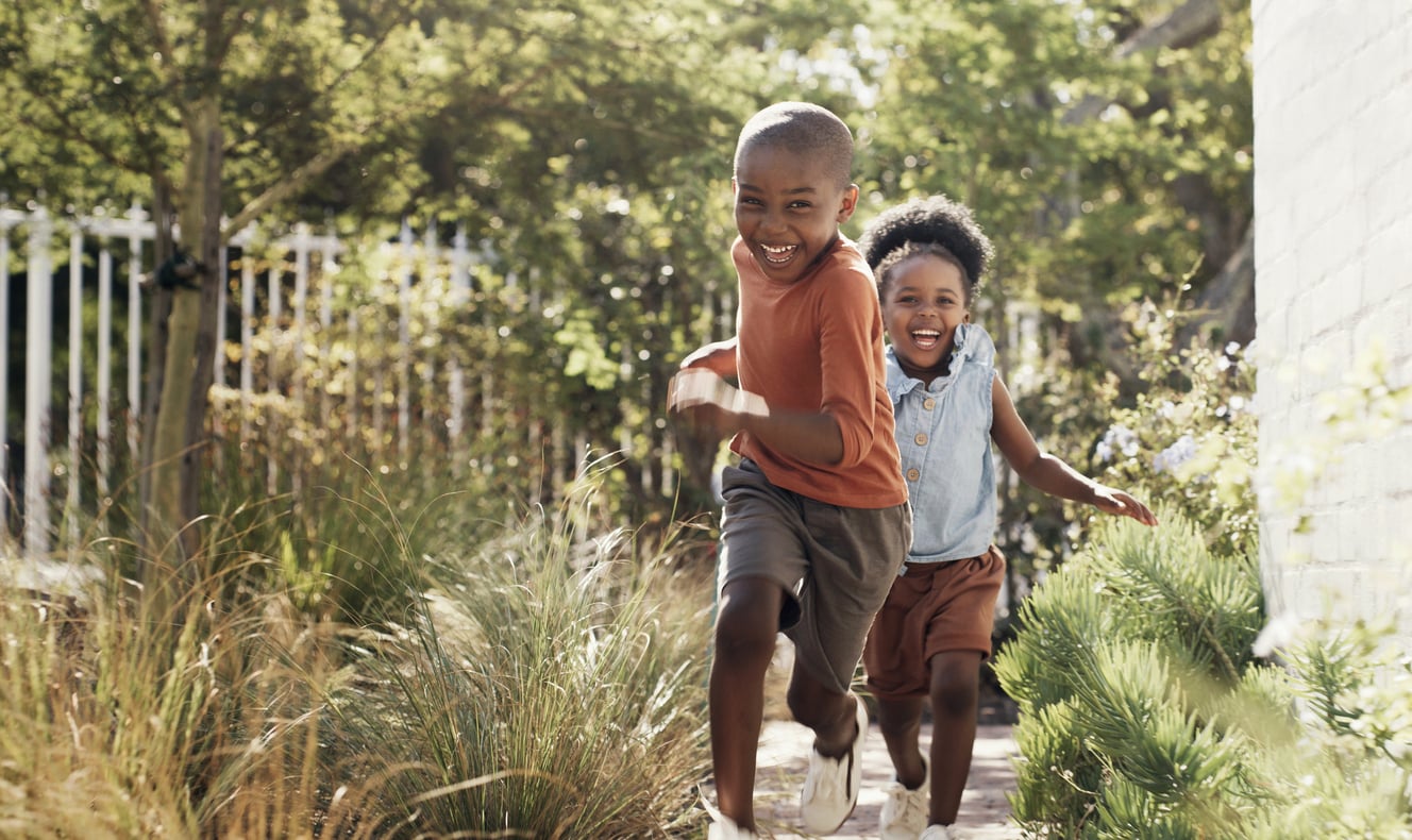 Two children run joyfully along a garden path, smiling and looking happy, surrounded by green plants and sunlight—capturing the simple pleasures of life before smartphones.