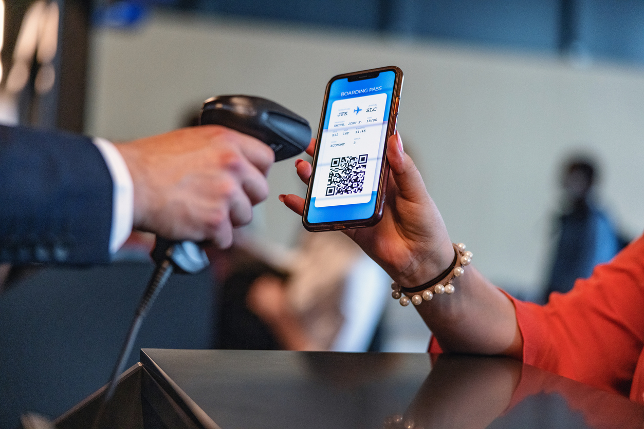 A person holds a smartphone displaying a boarding pass with a QR code while another person scans it with a handheld scanner at an airport check-in counter.