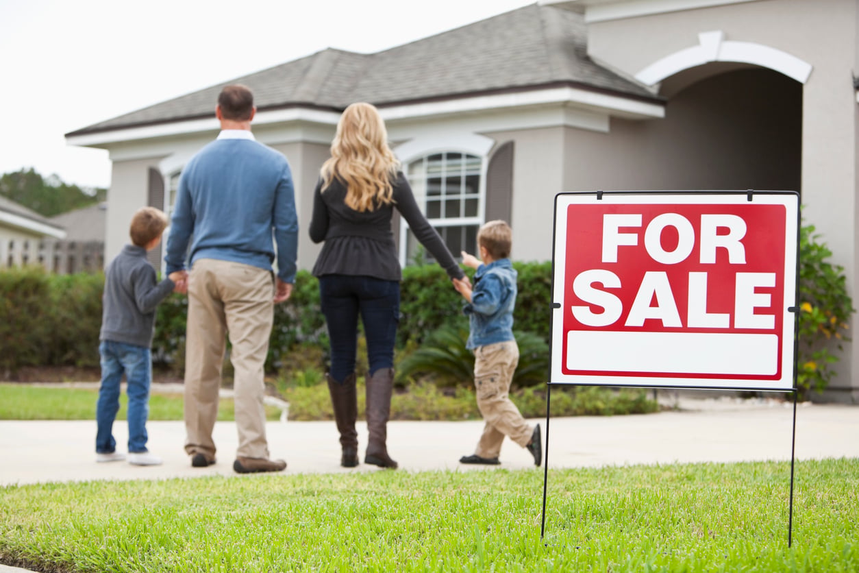 A family of four stands in front of a house, holding hands and looking at it. A large red and white “For Sale” sign is displayed on the lawn, reflecting how families in America are having fewer children today.