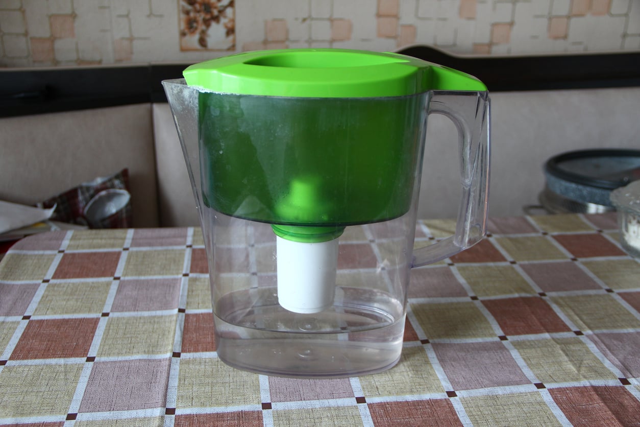 A transparent plastic water filter pitcher with a green lid and filter, one of the essential kitchen items, sits on a checkered tablecloth in a kitchen setting.