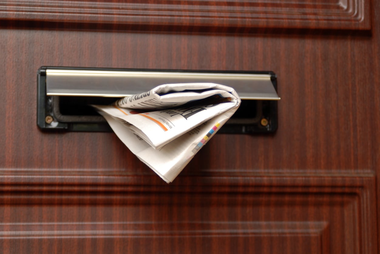 A folded newspaper, reminiscent of life before smartphones, partially sticks out of a mail slot in a wooden door.
