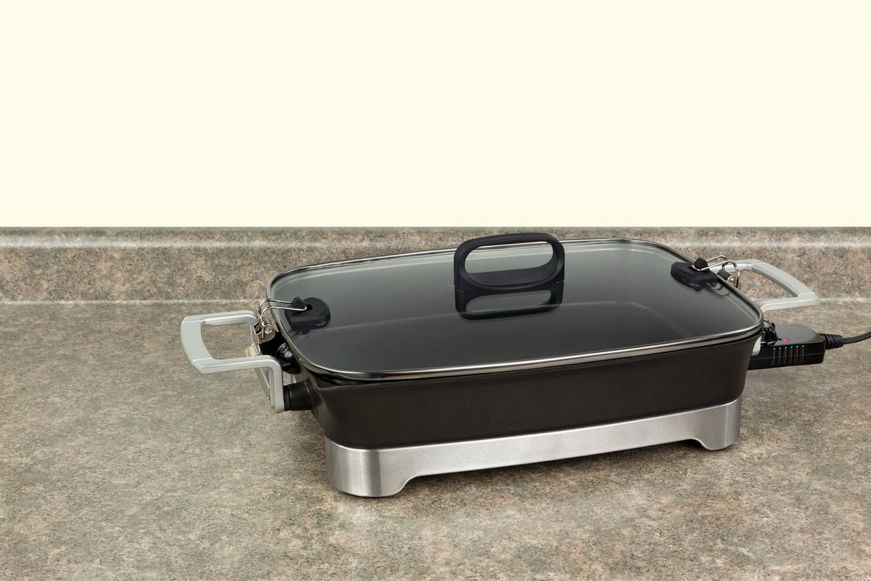 A black and silver electric skillet with a glass lid, one of the essential kitchen items, sits on a kitchen countertop, plugged in and ready for use.