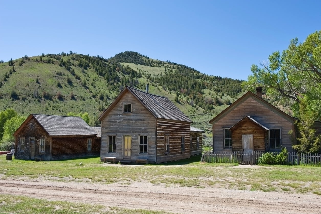 Three old wooden buildings stand on a grassy area with dirt roads in front, evoking the charm of historic American towns, set against a backdrop of green hills and a clear blue sky.
