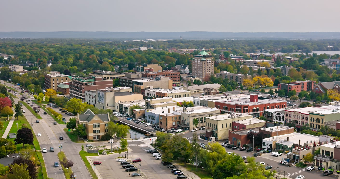 Aerial view of a small city with tree-lined streets, mid-rise buildings, parked cars, and a background of dense, green forest under a cloudy sky.