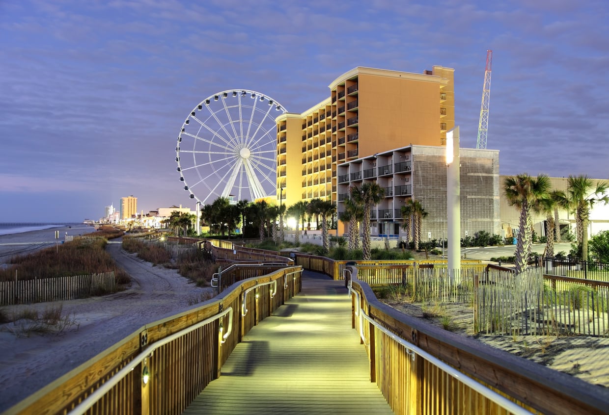 A wooden boardwalk leads to a beach with a large Ferris wheel, tall hotels, palm trees, and city lights under a cloudy evening sky—one of the many scenes that make this location stand out among affordable travel destinations.