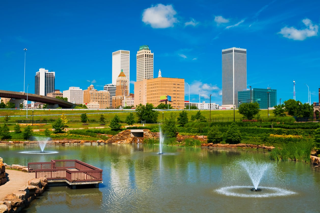 City skyline with tall buildings in background, blue sky above. In foreground, a pond with three fountains, greenery, and a small viewing deck—an inviting spot reminiscent of some of the world’s top 20 travel destinations.