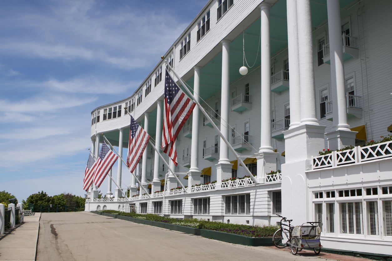 Large white hotel with tall columns and several American flags hanging at the front. Known as one of the area's affordable travel destinations, this charming spot features a bicycle parked near the entrance beneath a clear blue sky. Flowers line the walkway in front of the building, adding to its inviting atmosphere.