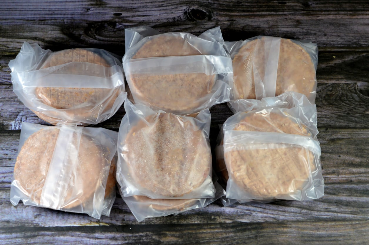Six frozen hamburger patties individually wrapped in plastic, arranged in two rows of three, on a wooden surface.