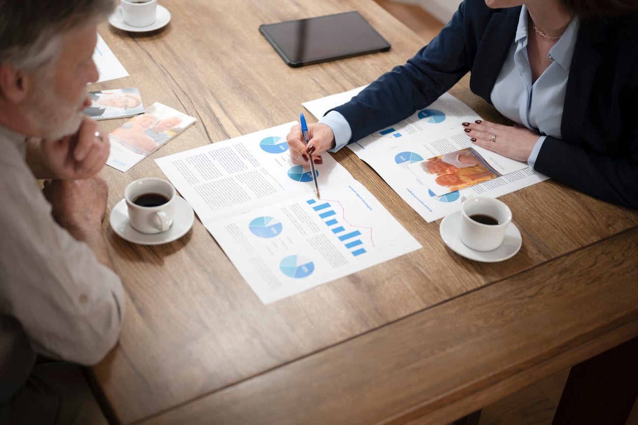 Two people sit at a wooden table with coffee cups, discussing printed charts and graphs. As one person points at a blue bar graph with a pen, the conversation centers on how Women Are Changing Their Spending Behaviors. A tablet and additional papers are also on the table.