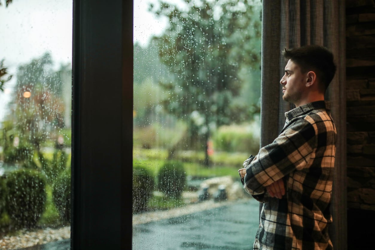 A man in a plaid shirt stands indoors with arms crossed, gazing thoughtfully out a large window dotted with raindrops, while a rainy, blurred landscape is visible outside.