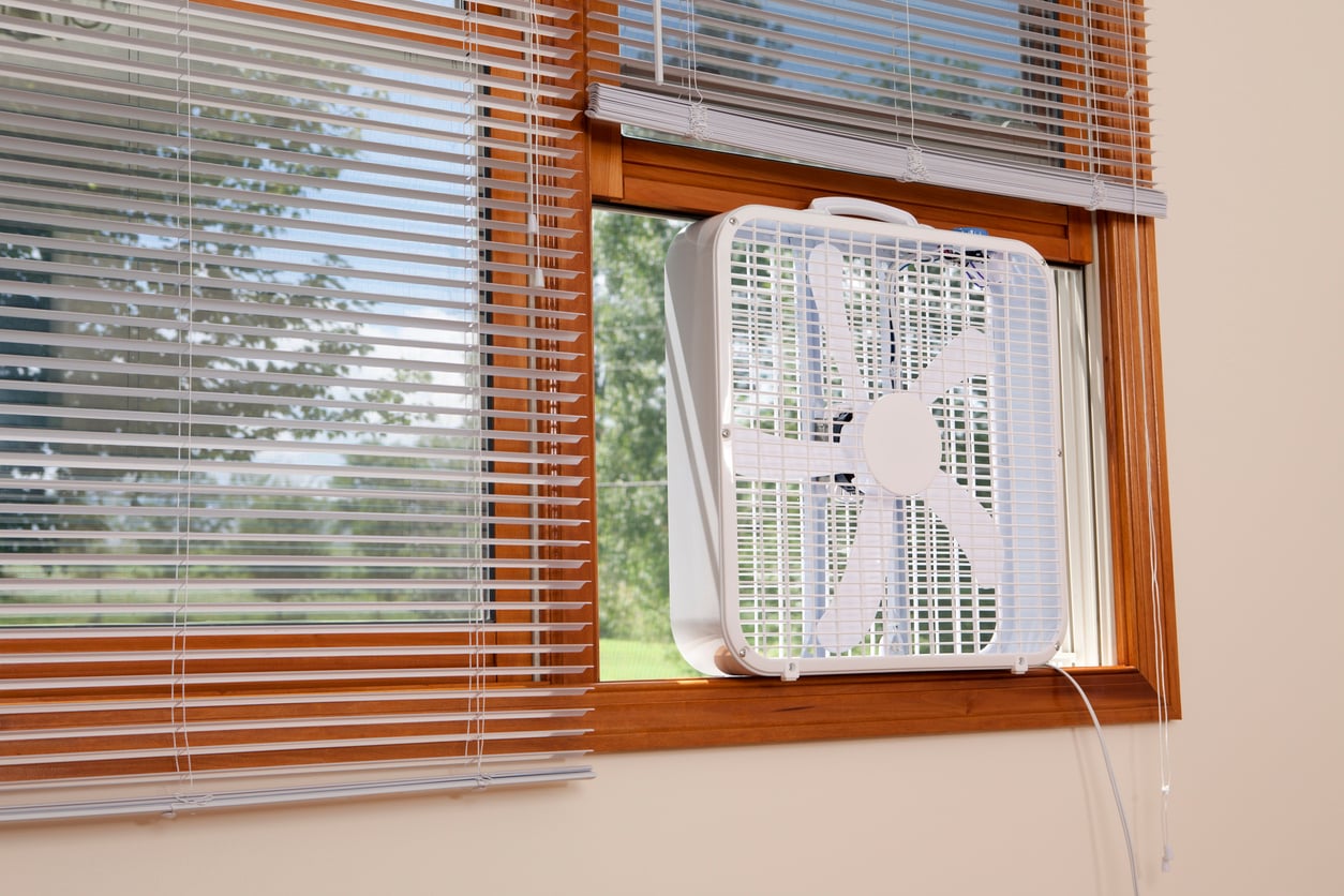 A white box fan is placed in an open window framed with wooden trim and partially covered by horizontal blinds. Trees and grass are visible outside through the window.
