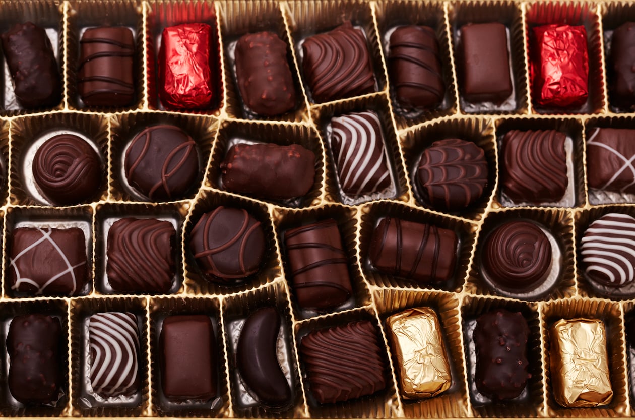A box of assorted chocolates arranged in rows, featuring various designs, shapes, and foil-wrapped pieces in red and gold among the unwrapped chocolates.