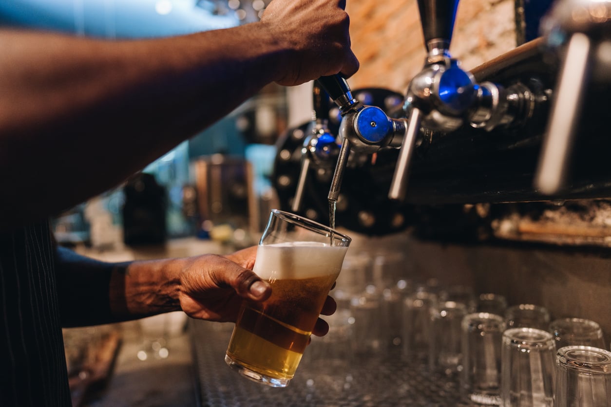 A person fills a glass with draft beer from a tap at a bar, with several empty glasses lined up in the background.
