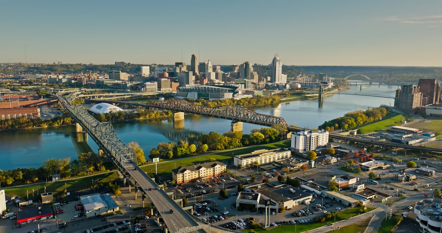 Aerial view of a travel-friendly city skyline with multiple bridges crossing a river, urban buildings, trees, and roads, under a clear sky during daylight.