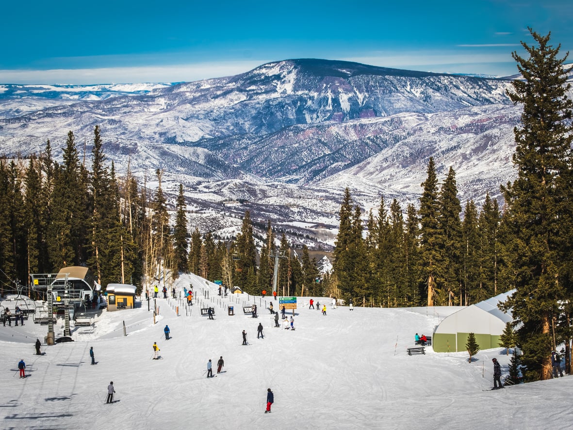 Skiers and snowboarders glide down a snowy mountain slope lined with pine trees, with chairlifts, buildings, and distant snow-covered mountains under a clear blue sky.