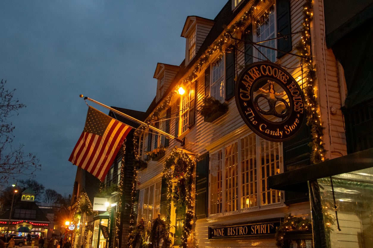 A warmly lit street scene at dusk shows a decorated building with an American flag and a sign reading “Clarke Cooke House: The Candy Store,” adorned with string lights and greenery—capturing the festive charm that makes it one of the best Christmas towns in America.