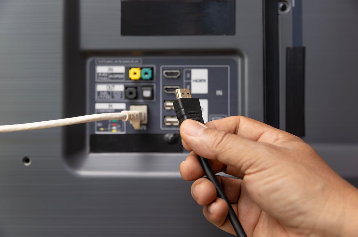 A hand holds an HDMI cable near the HDMI port on the back of a TV, next to an Ethernet cable that is already plugged in. Various input ports and labels are visible on the television, serving as a reminder to carefully choose gadgets and avoid waste of money products.
