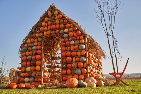 A small halloween house made of stacked pumpkins with a straw roof stands on grass. A red chair sits nearby, and the sky is clear and blue.