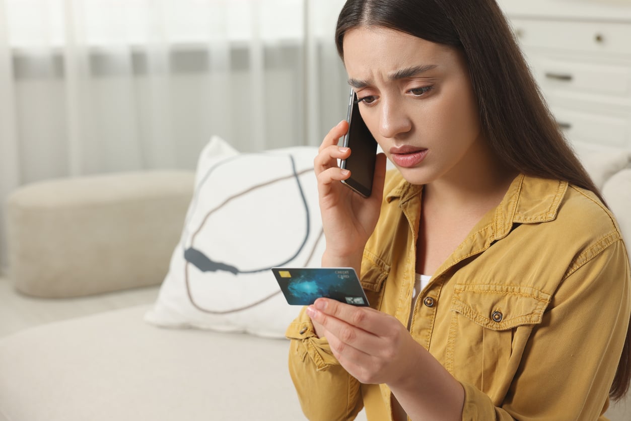 A woman in a yellow shirt sits on a couch, looking worried while talking on the phone and holding a credit card, possibly dealing with an issue related to her monthly bills.